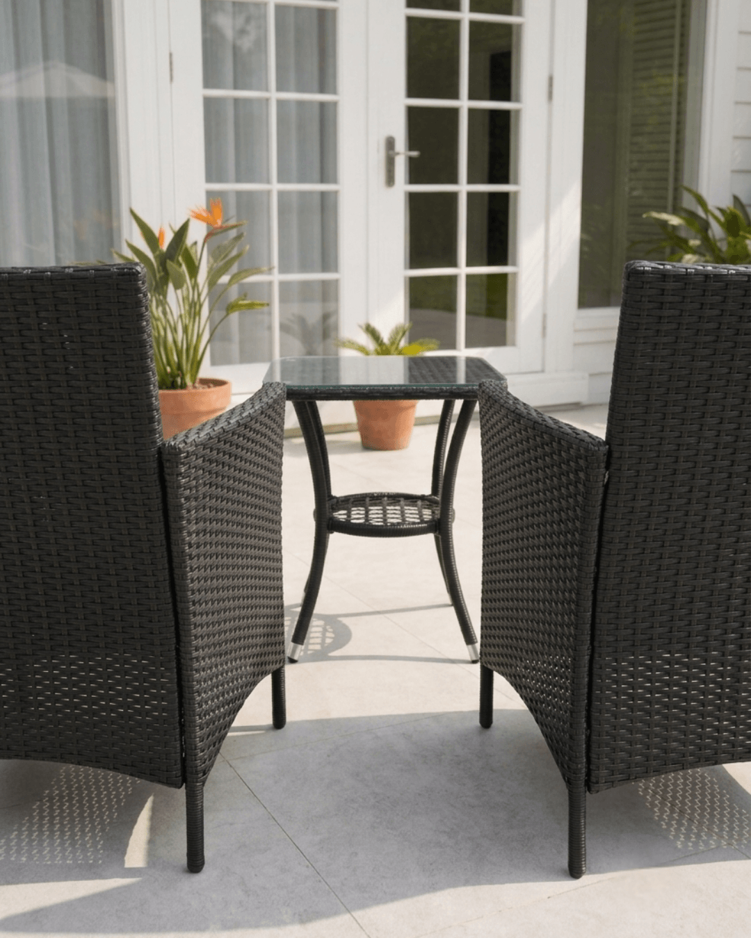 A 3-piece outdoor patio dining set featuring two black woven wicker armchairs and a matching square glass-top side table, positioned on a light gray tiled patio in front of white French doors