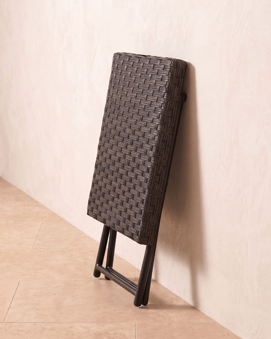 A collapsible brown small rattan garden table leaning against the wall