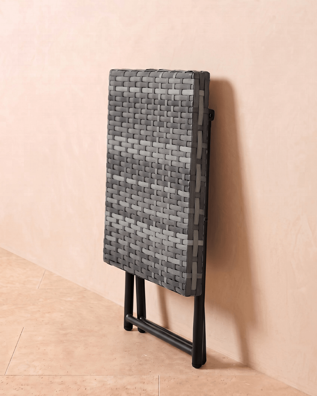 A grey rattan small garden table folded and leaning against the wall