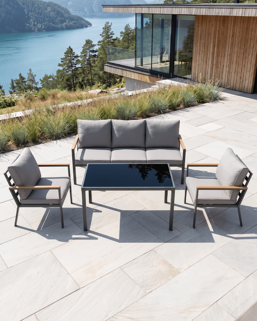 A 4-piece outdoor patio furniture set including a three-seater sofa, two armchairs, and a rectangular dining table, arranged on a light stone patio overlooking a scenic lake and forest view.