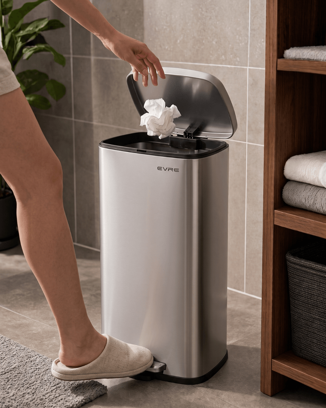 A stainless steel bathroom bin shown in a bathroom setting with a person's leg and foot stepping on the pedal and a tissue being thrown into the bin
