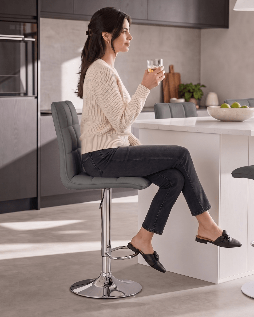 A woman sitting on a grey upholstered bar chair with a chrome base in a modern kitchen