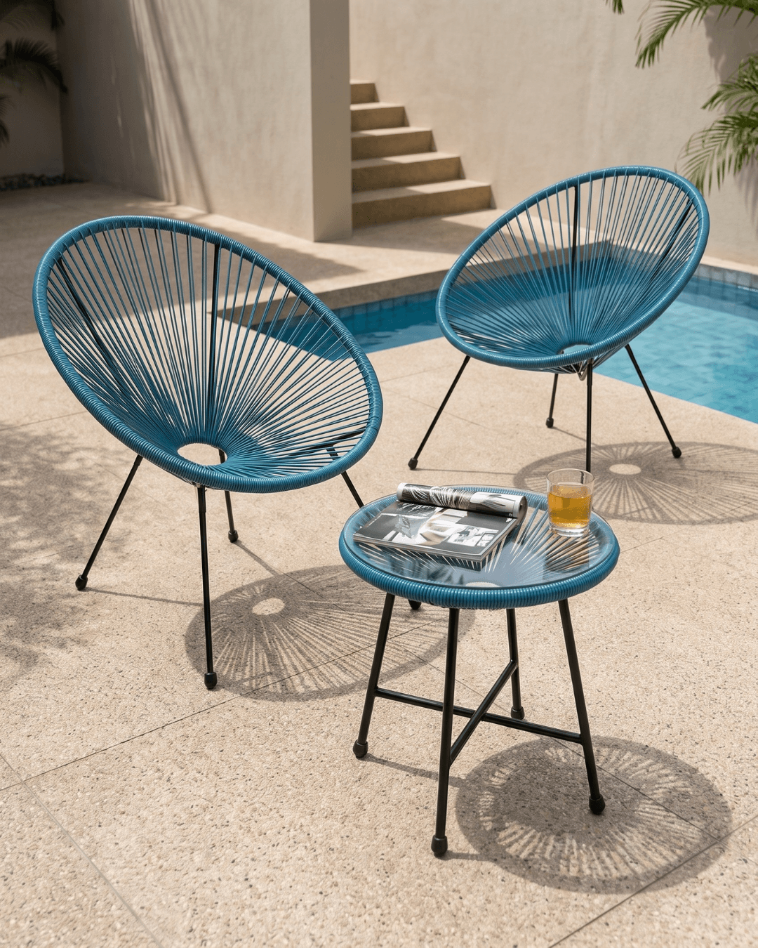 Two teal blue woven accent chairs with black metal legs are arranged on a stone patio beside a swimming pool, with a small matching side table in the foreground holding a magazine and a glass of orange juice
