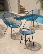 Two teal blue woven accent chairs with black metal legs are arranged on a stone patio beside a swimming pool, with a small matching side table in the foreground holding a magazine and a glass of orange juice