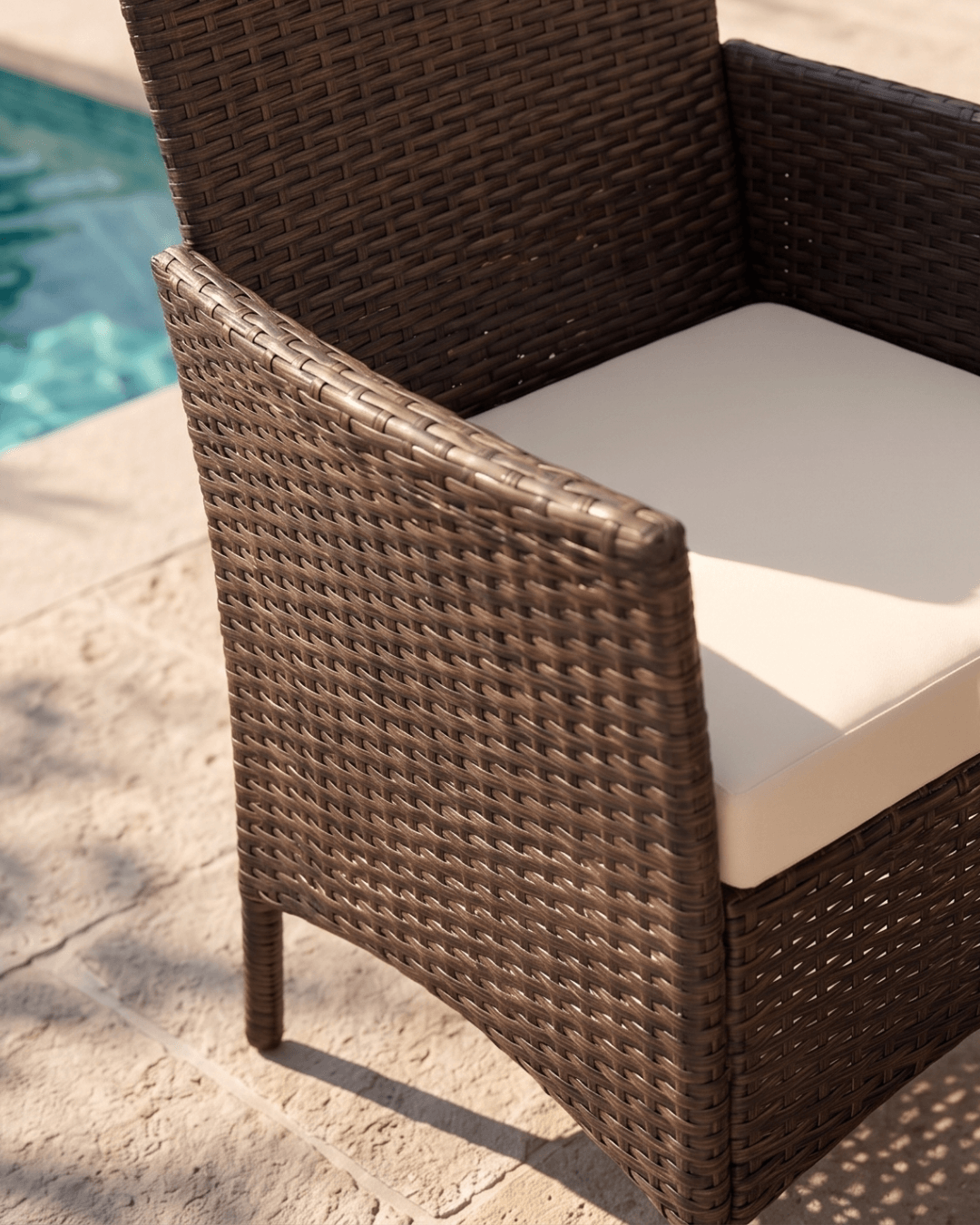 A close-up of a brown woven wicker patio chair with a cream-colored seat cushion, positioned outdoors on a stone patio
