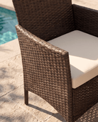 A close-up of a brown woven wicker patio chair with a cream-colored seat cushion, positioned outdoors on a stone patio