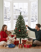 A family of three decorates a small artificial Christmas tree with red and gold ornaments in a bright living room with a snowy window view. Gift boxes are wrapped around the base of the tree.