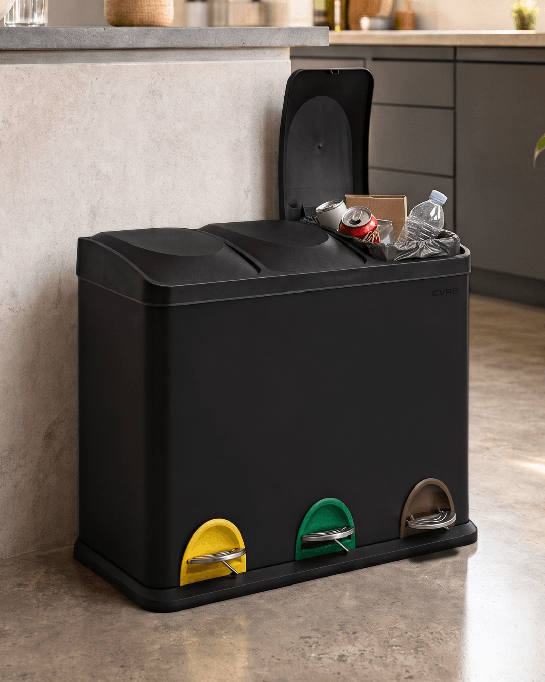 A black triple compartment trash bin with a hinged lid open, set in a kitchen next to a counter. The bin features three coloured foot pockets at the bottom: yellow, green, and brown.