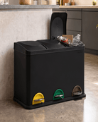 A black triple compartment trash bin with a hinged lid open, set in a kitchen next to a counter. The bin features three coloured foot pockets at the bottom: yellow, green, and brown.
