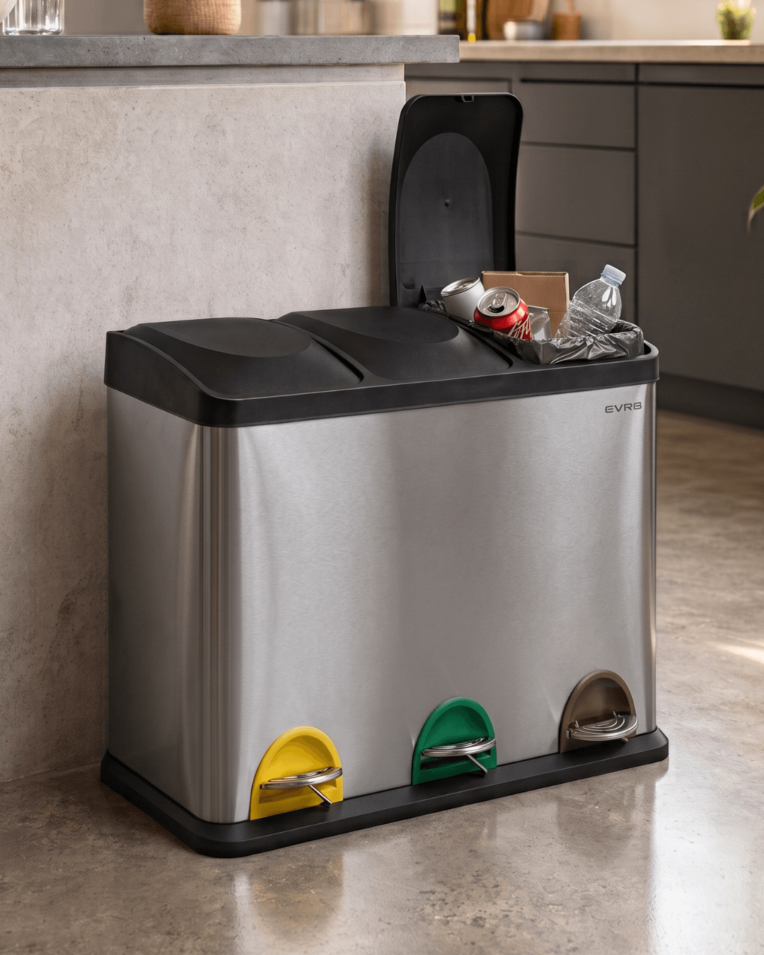 A stainless steel three-compartment waste bin with black lids and coloured foot pedals, shown in a kitchen setting with trash inside