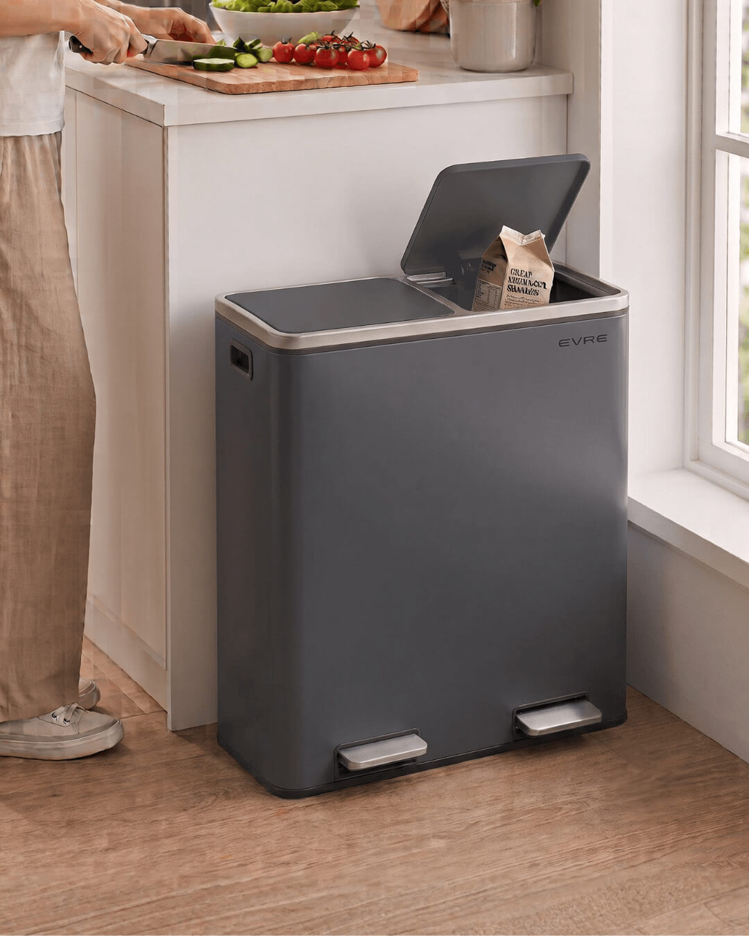 A large gray kitchen pedal bin with an open lid, placed on a wooden floor next to a kitchen counter