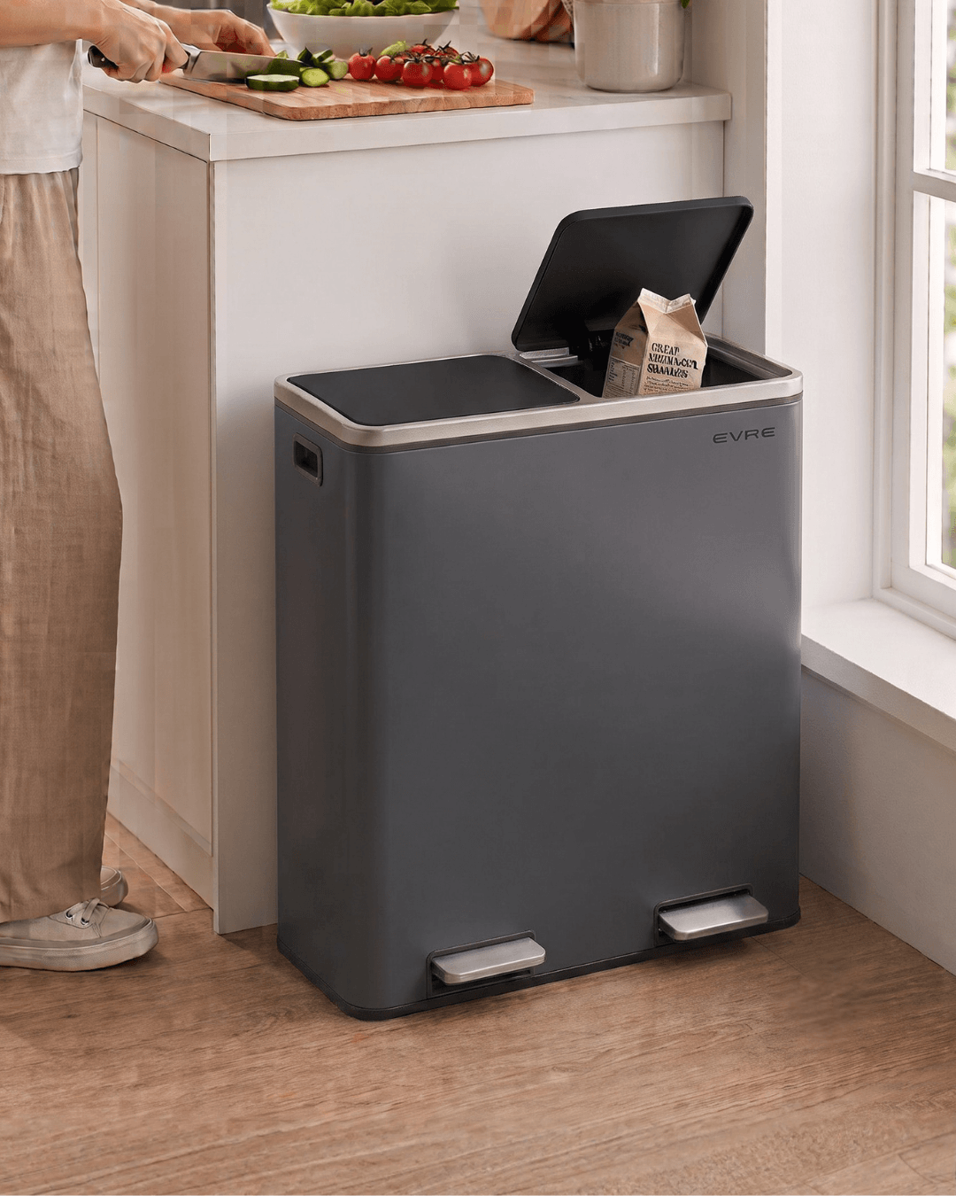 A dark gray dual-compartment kitchen bin with a hinged lid positioned next to a kitchen counter 