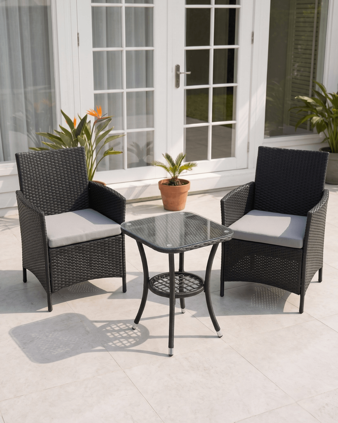 A 3-piece outdoor patio furniture set featuring two black woven wicker armchairs with gray cushions and a matching square glass-top side table, arranged on a light stone patio in front of white French doors