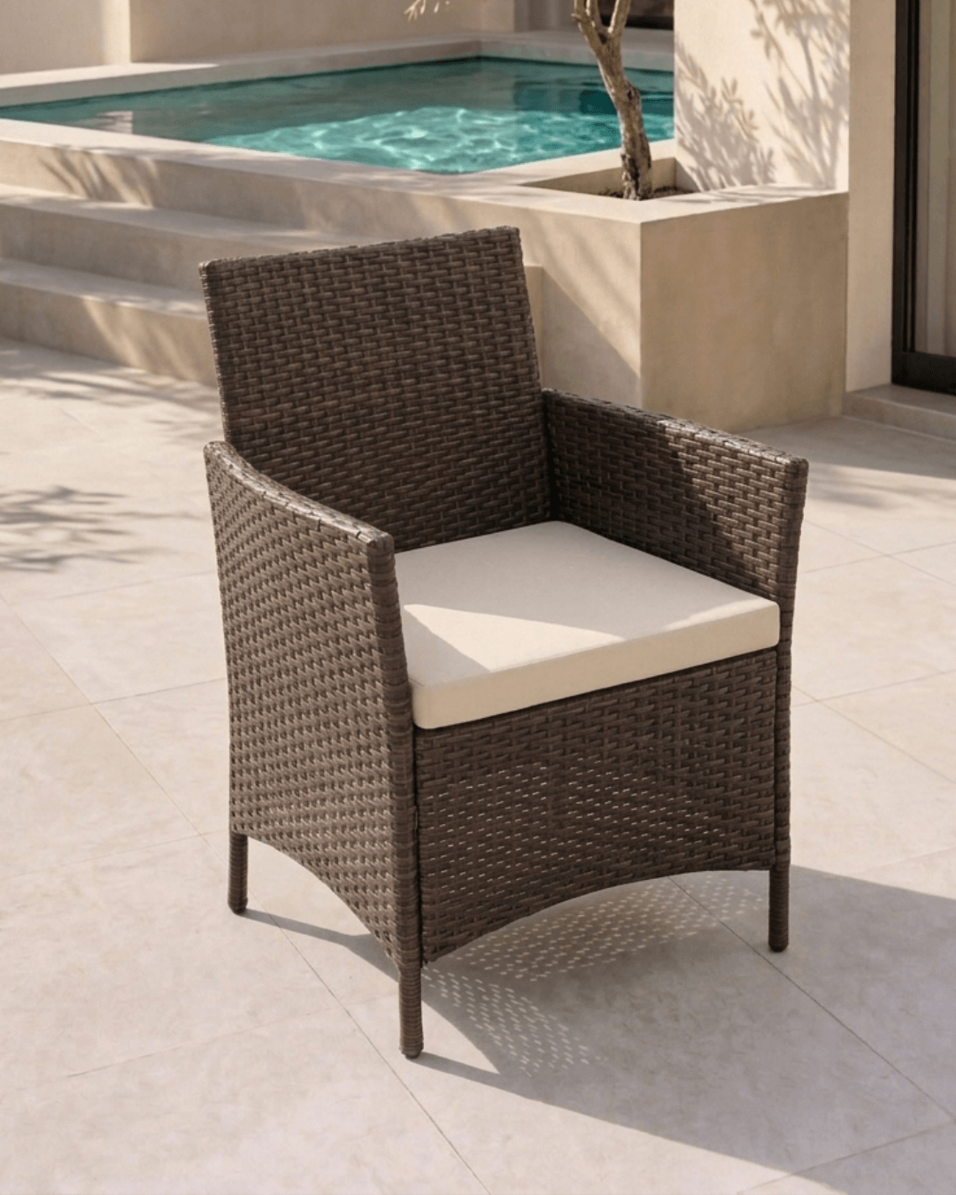 A brown woven outdoor patio armchair with a cream-colored seat cushion, positioned on a light stone tiled patio next to a swimming pool