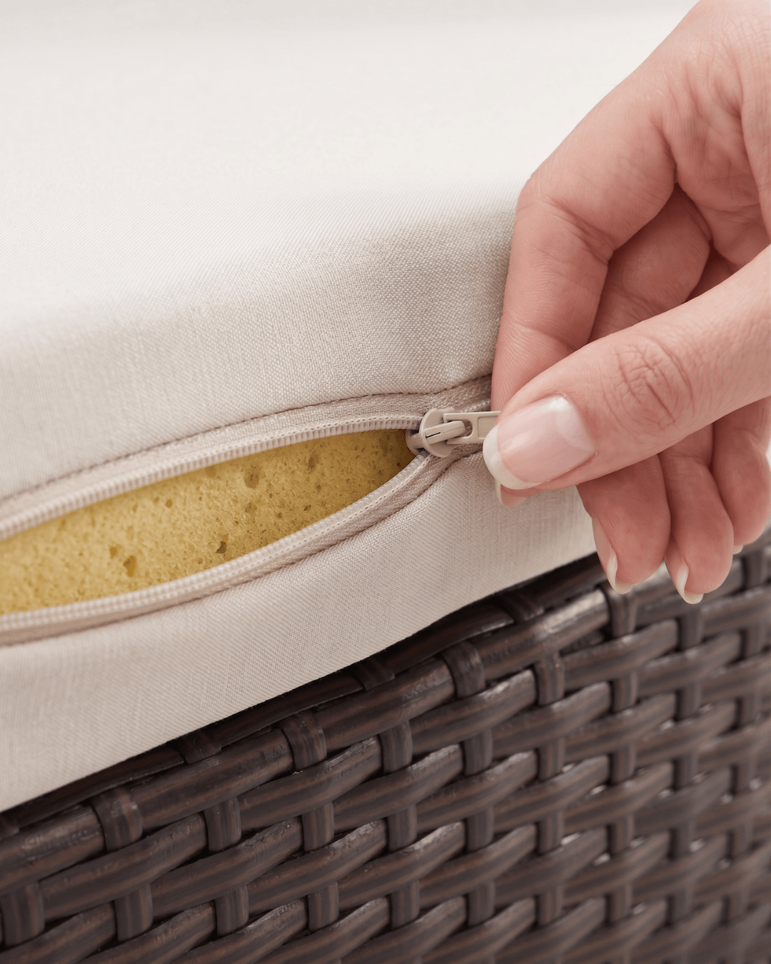 Close-up of a hand unzipping a cream outdoor cushion over a dark brown woven wicker surface, showing the yellow sponge insert