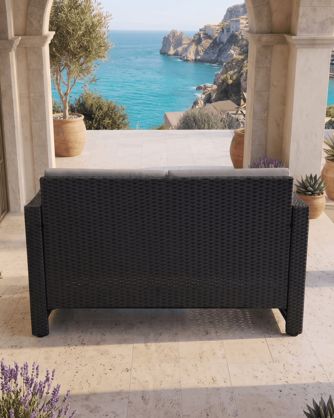 A black woven outdoor loveseat sofa viewed from the back, positioned on a stone terrace with a scenic view of a turquoise sea and rocky coastline in the background