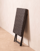 A collapsible brown small rattan garden table leaning against the wall