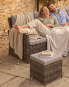 A couple relax on a dark brown wicker outdoor lounge sofa with grey cushions, draped with cream throw blankets