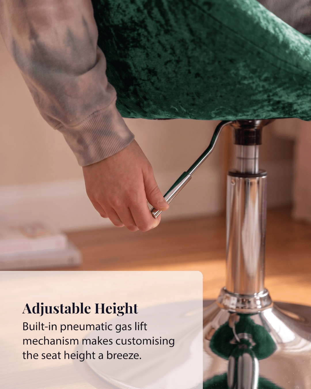 A close-up of a person's hand adjusting the chrome lever of a dark green velvet upholstered stool with a built-in pneumatic gas lift mechanism
