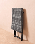A grey rattan small garden table folded and leaning against the wall