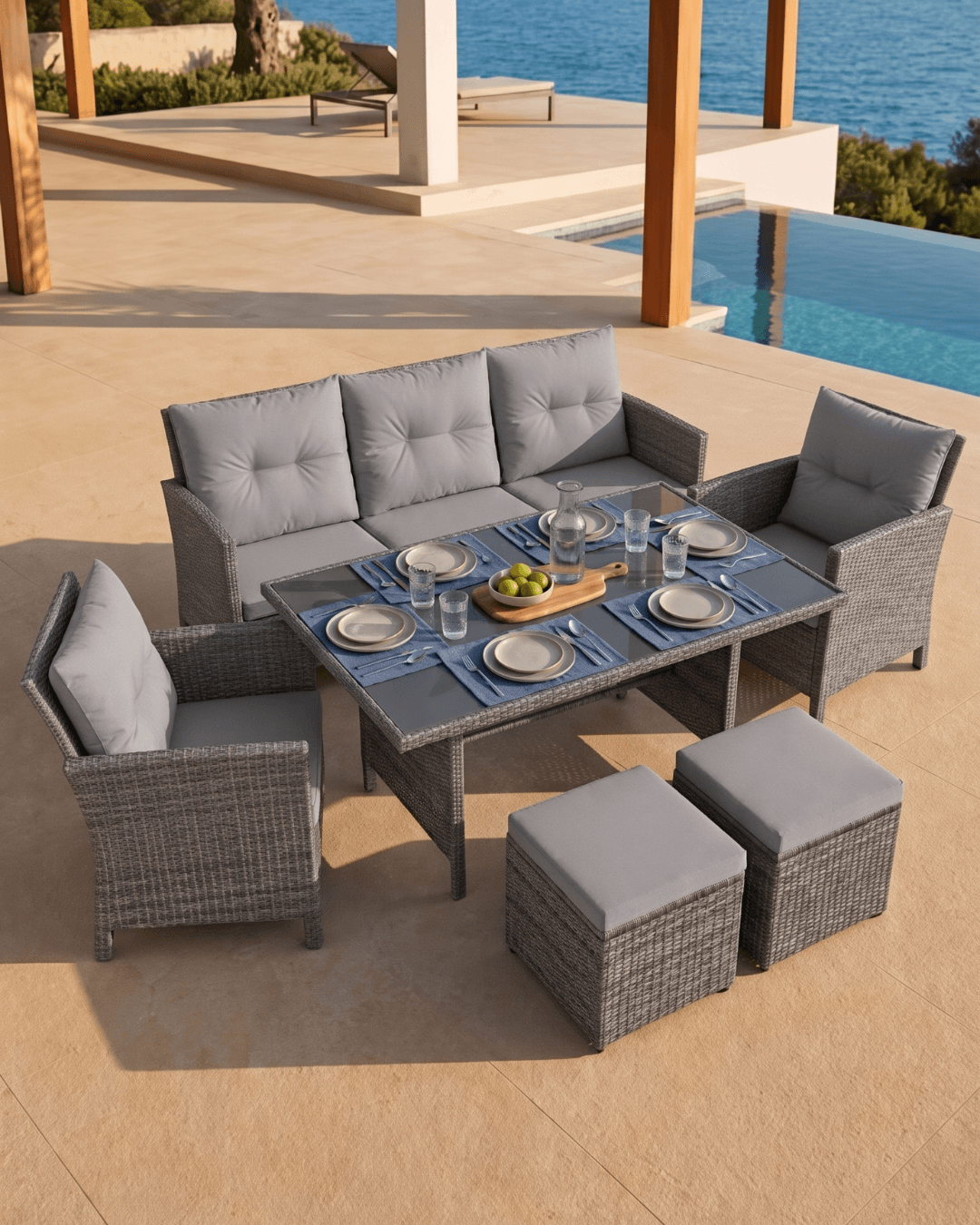 Gray wicker outdoor patio dining set with a three-seat sofa, two armchairs, a rectangular coffee table, and two ottomans, arranged on a beige stone patio near a pool and ocean view