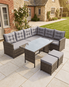 Rattan corner sofa garden set including wicker sectional sofas with grey cushions, a matching rectangular dining table, and two ottomans, shown on a stone patio in a British garden