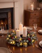 Pre-lit frosted wreath ringing three white pillar candles as a Christmas table decoration with pine cones and red berries