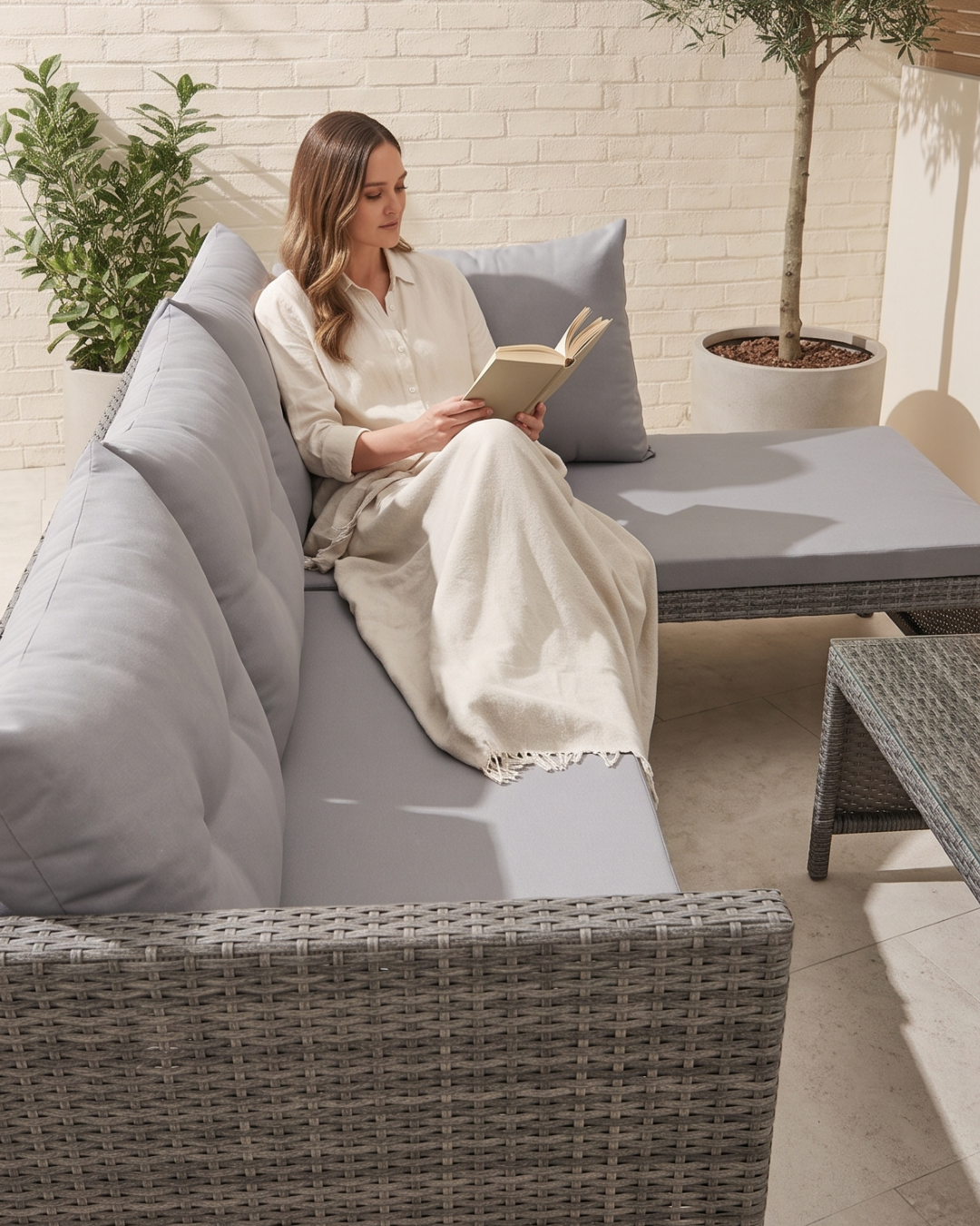 A woman lounging on a grey rattan sofa lounger wrapped in a cream blanket and reading a book