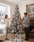 A couple decorating a 7ft large snowy Christmas tree decorated with ornaments, a star topper, and warm string lights in a cosy living room