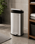 Stainless steel pedal bin with lid on tiled bathroom floor next to a wooden shelf with folded towels