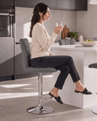 A woman sitting on a grey upholstered bar chair with a chrome base in a modern kitchen