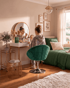 A cozy bedroom scene with a young girl sitting in a green velvet swivel chair, looking into a round mirror at a vanity table